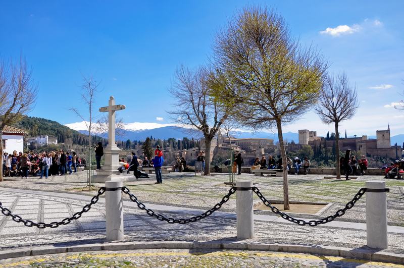 Le mirador de San Nicolás, dans l’Albaicín, avec sa croix de pierre, des gens accoudés et l’Alhambra et la sierra au fond